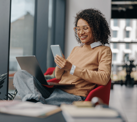 Woman Sitting on a Chair Smiling Looking at Phone with Laptop