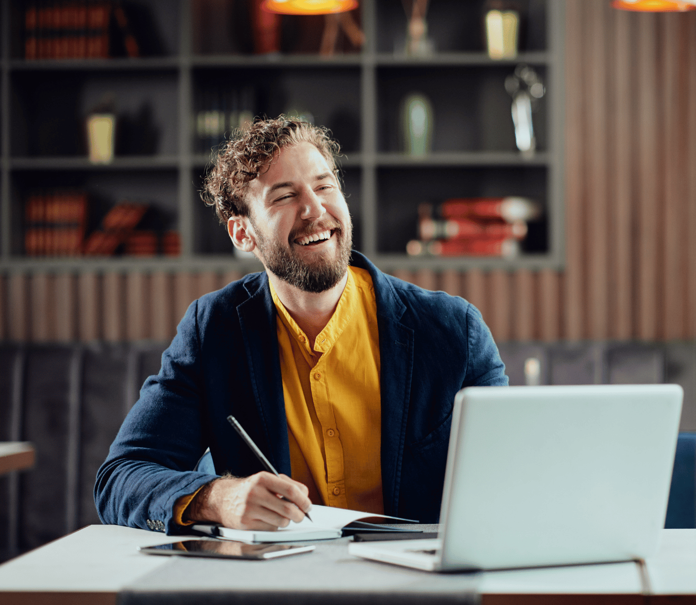 A hip man wearing a yellow shirt taking notes on laptop while laughing.